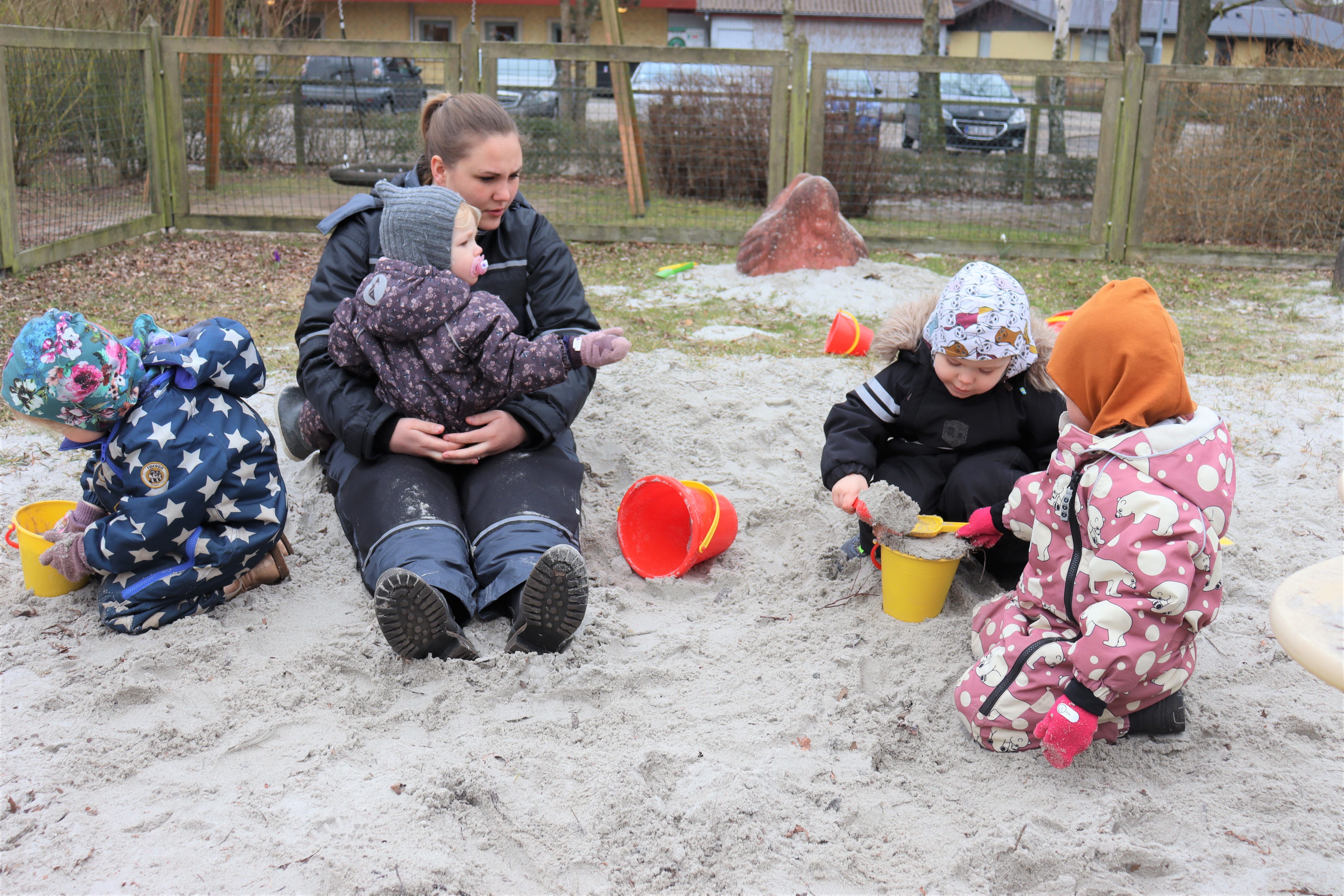 Pædagog sidder i sandkassen med flere børn, omkring sig.